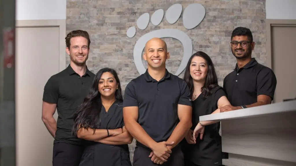 Five people in black shirts stand smiling in a modern office with a stone wall and a large foot logo behind them. Three are standing, two are beside a reception desk, creating a welcoming atmosphere.