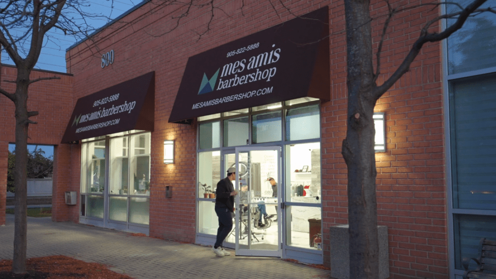 A person stands outside the glass door entrance of Mes Amis Barbershop in a brick building at dusk, with the interior lights on and the shop name visible on the awning above.