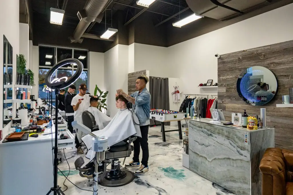 Modern barbershop interior with barbers cutting clients’ hair. Two barbers work on customers seated in chairs, while two other people stand by the window. The space is bright, stylish, and decorated with modern furniture.