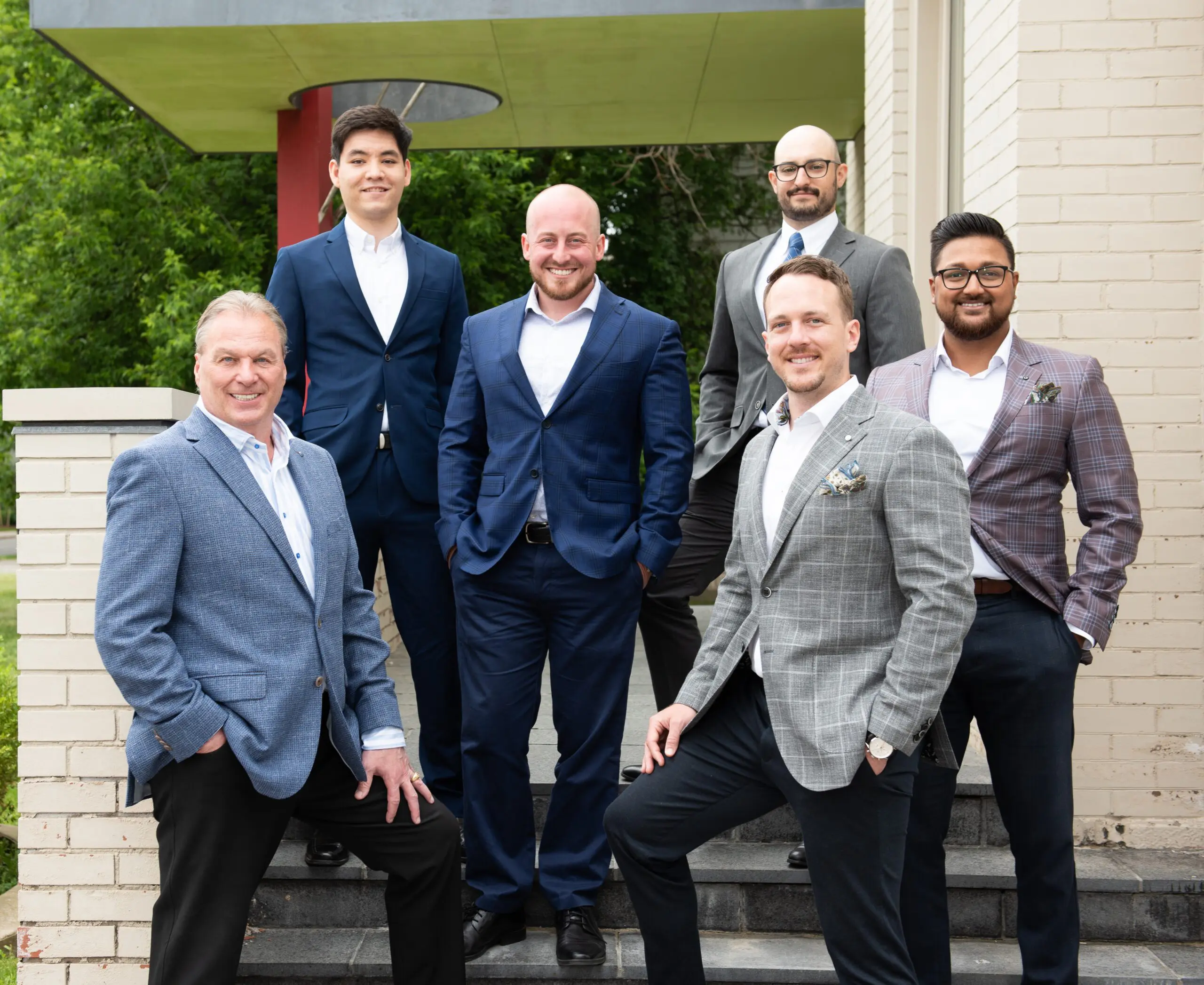 Six men in business suits pose confidently on outdoor steps in front of a brick building and greenery, smiling for the camera.
