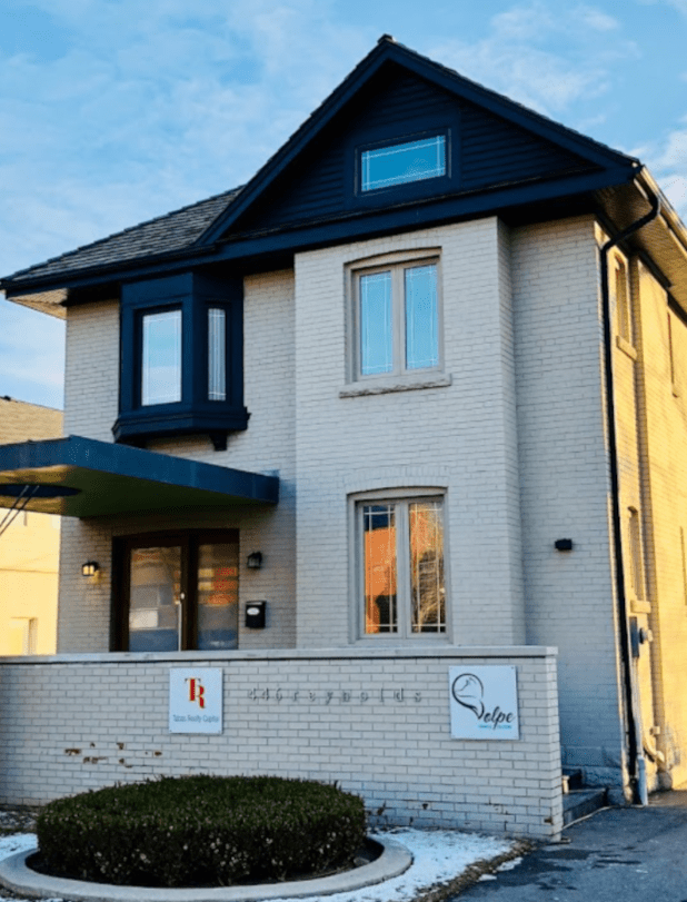 A two-story beige brick building with a dark roof and bay window, featuring office signs for Riz Realty Group and Velpe near the entrance, and a small circular hedge in front.