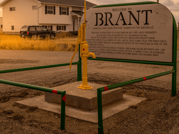 A metal sign reading Welcome to Brant stands beside a yellow water pump on a concrete base, enclosed by green and red railings. A house and parked vehicle are visible in the background.