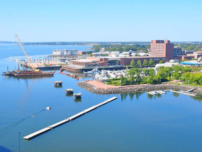 Aerial view of a harbor with boats docked at piers, a crane on the waterfront, red-brick buildings, green trees, and calm blue water under a clear sky.