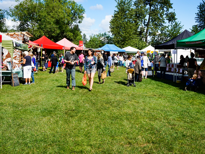 People walk and shop among colorful tents at an outdoor farmers market on a sunny day, surrounded by green grass and trees. Some carry bags, while vendors display goods at their stalls.