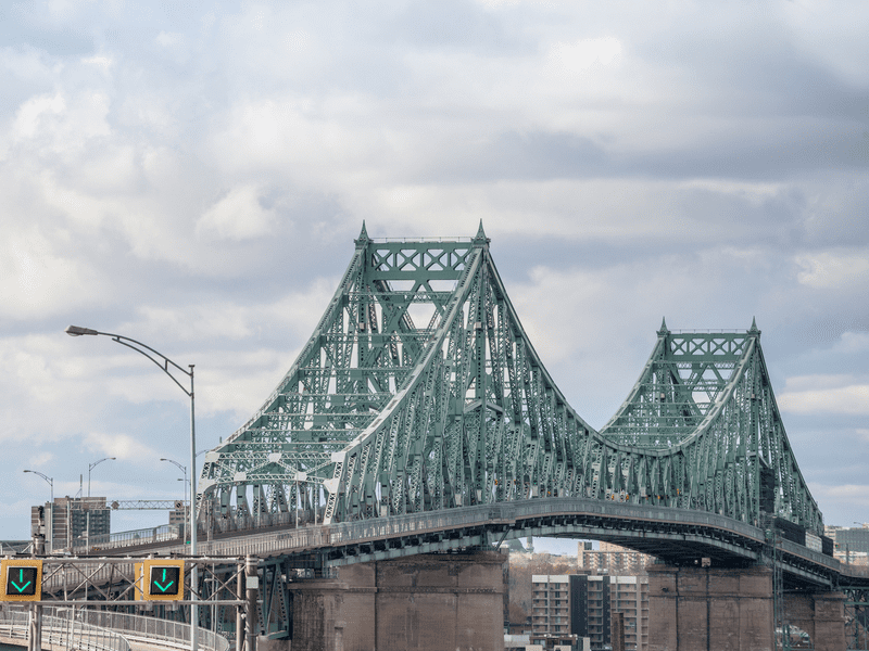 A large green steel truss bridge with two peaks stretches across a river, connecting city buildings under a cloudy sky. Traffic signs with green arrows are visible at the entrance.