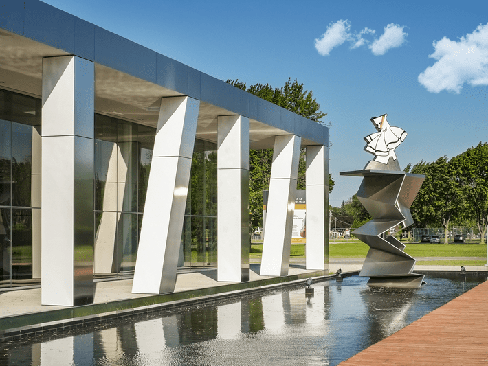 A modern building with large, white, slanted columns stands next to a reflecting pool featuring a geometric metal sculpture topped with a stylized figure. Green trees and blue sky with clouds are in the background.