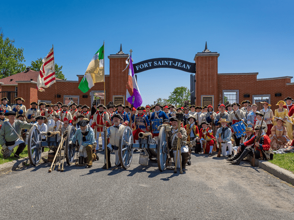 A large group of people in historical military costumes pose with cannons and flags in front of the entrance gate reading Fort Saint-Jean on a sunny day.