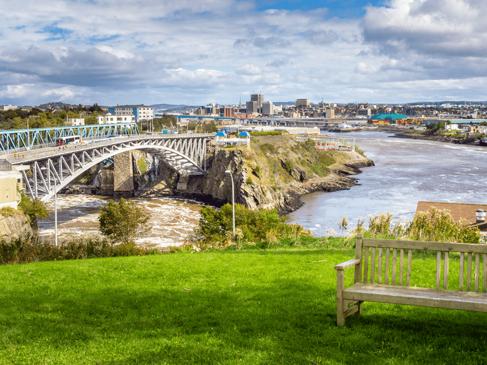 A wooden bench sits on green grass overlooking a river with a metal bridge and rapids; a cityscape with buildings is visible in the background under a partly cloudy sky.