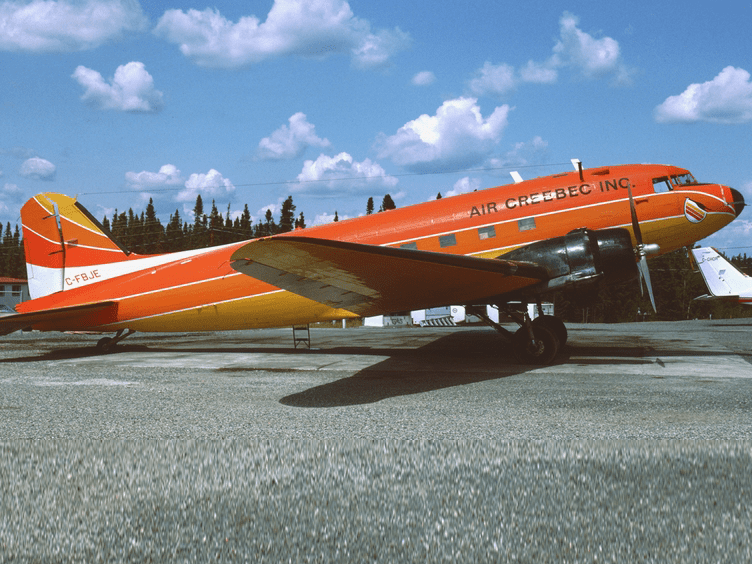 A bright orange and yellow Air Creebec Inc. propeller plane is parked on a tarmac under a blue sky with scattered clouds, with trees in the background.