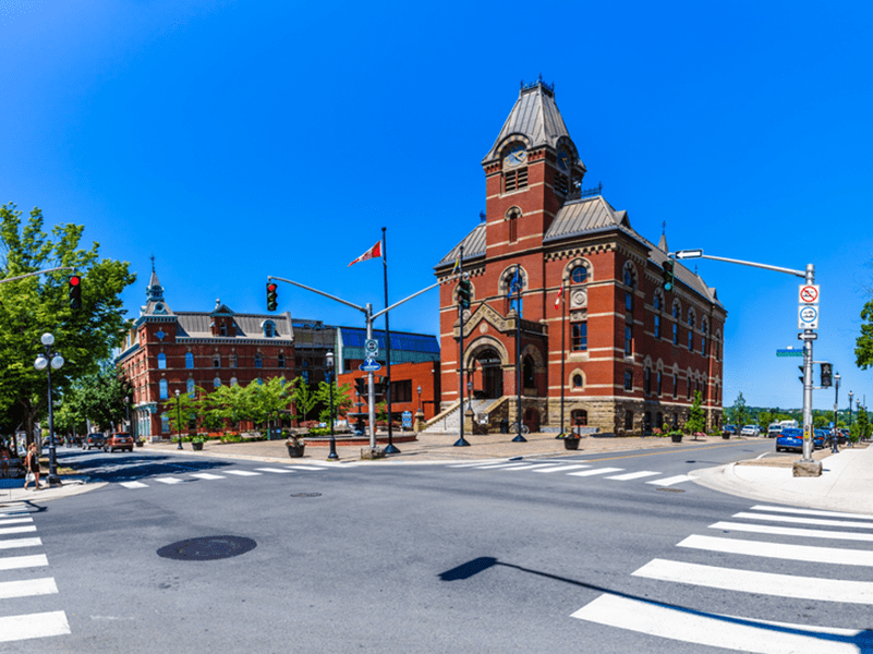 A historic red-brick building with a clock tower stands at a street corner on a sunny day, surrounded by crosswalks, traffic lights, and a clear blue sky.